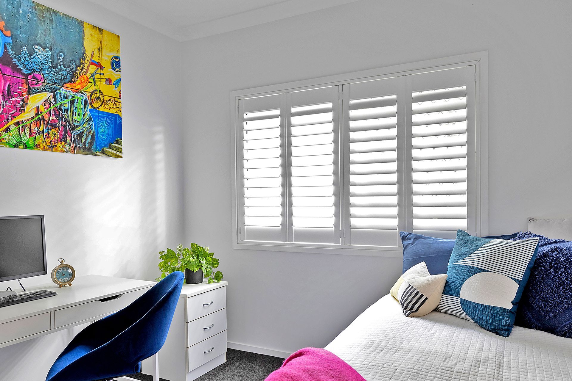 Bedroom with desk, bed, and window with shutters. Colorful art on wall.