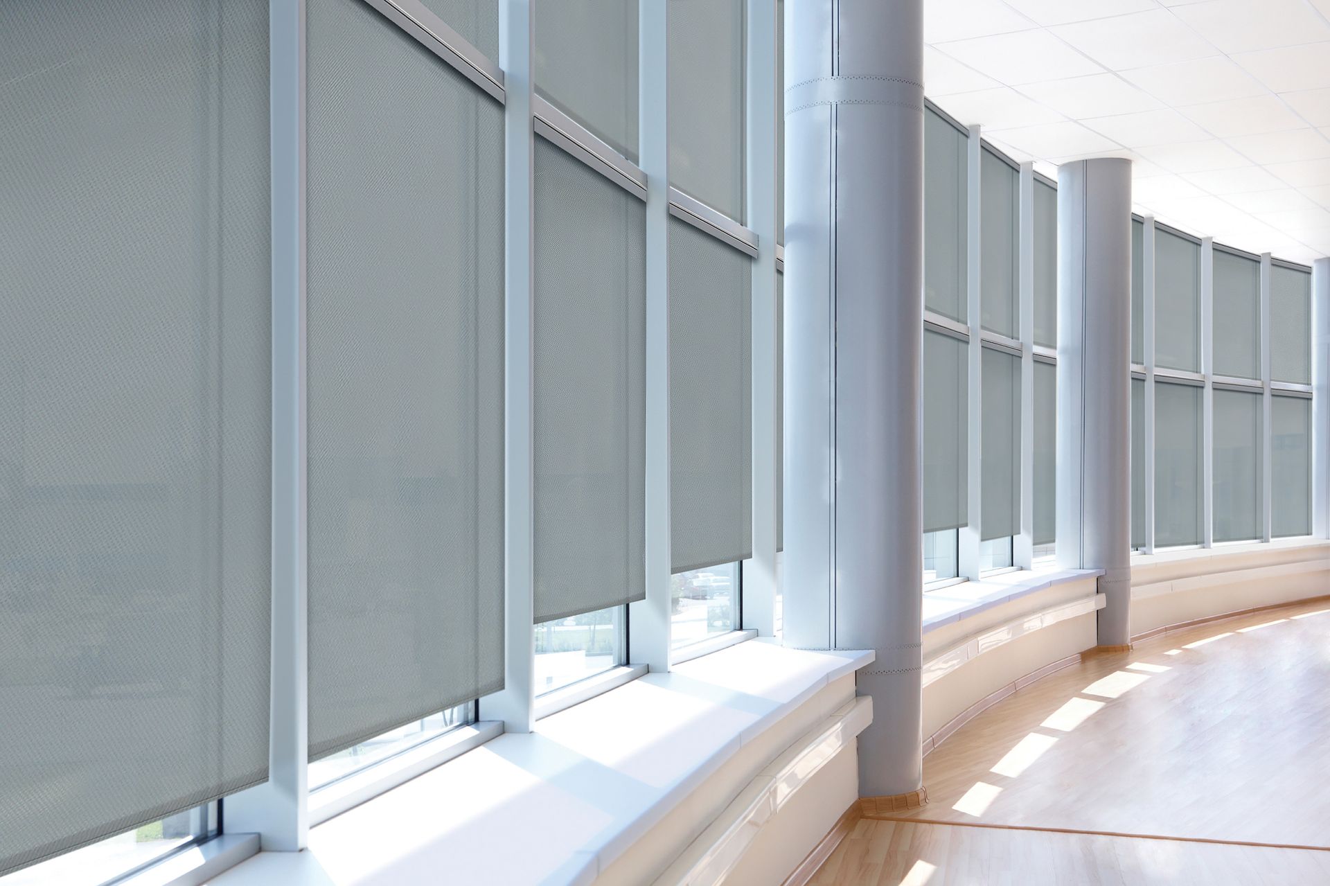 Row of light gray roller shades in a building with large windows and white support columns.