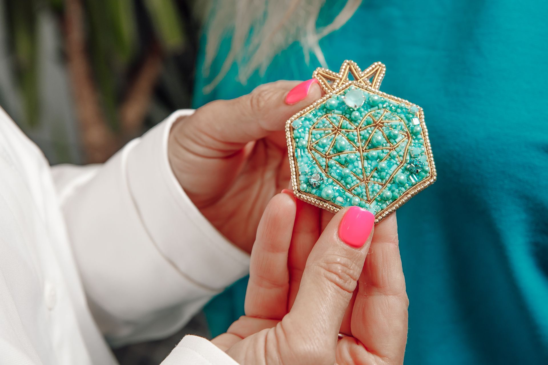 Person holding a turquoise beaded pomegranate brooch with gold trim and a pink manicure.
