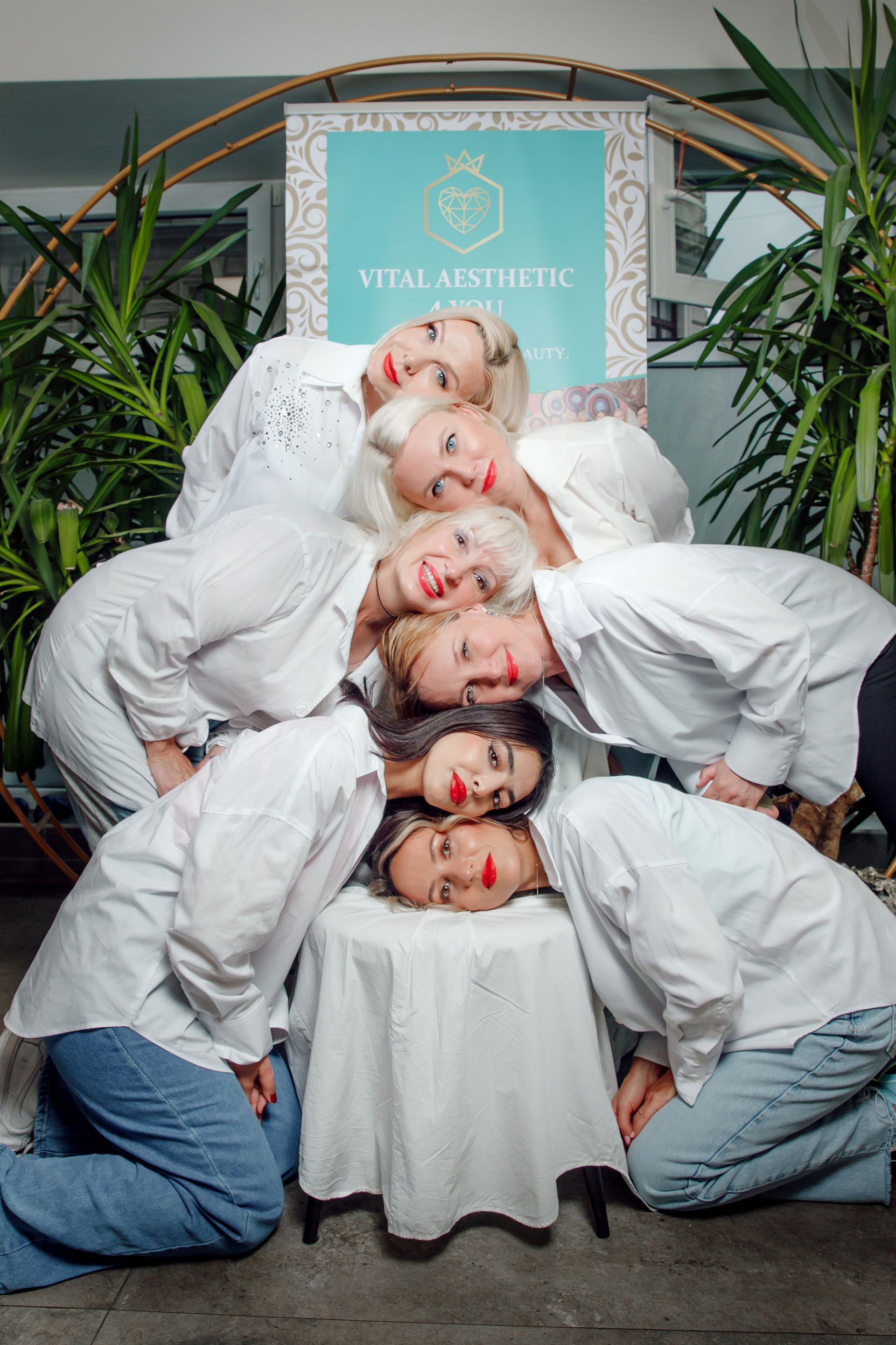 Seven women in white shirts and jeans huddle around a table. A sign that reads “Vital Aesthetic” is visible.