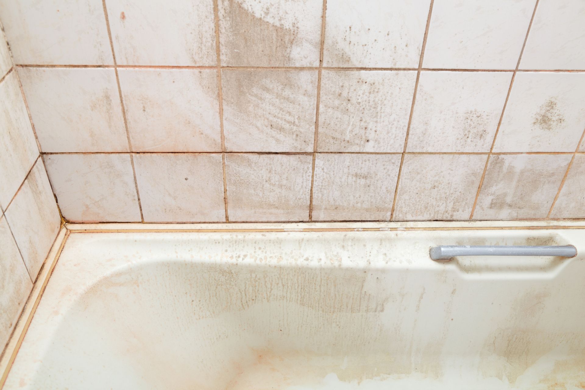 A bathtub in a bathroom with dirty tiles and mold on the wall.