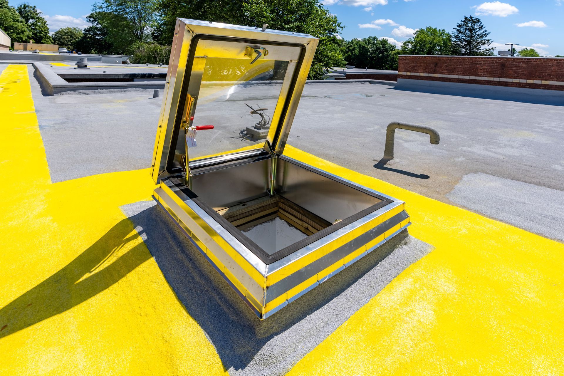 Open, metal roof hatch on a yellow painted section of a flat roof, with trees and a blue sky in the background.