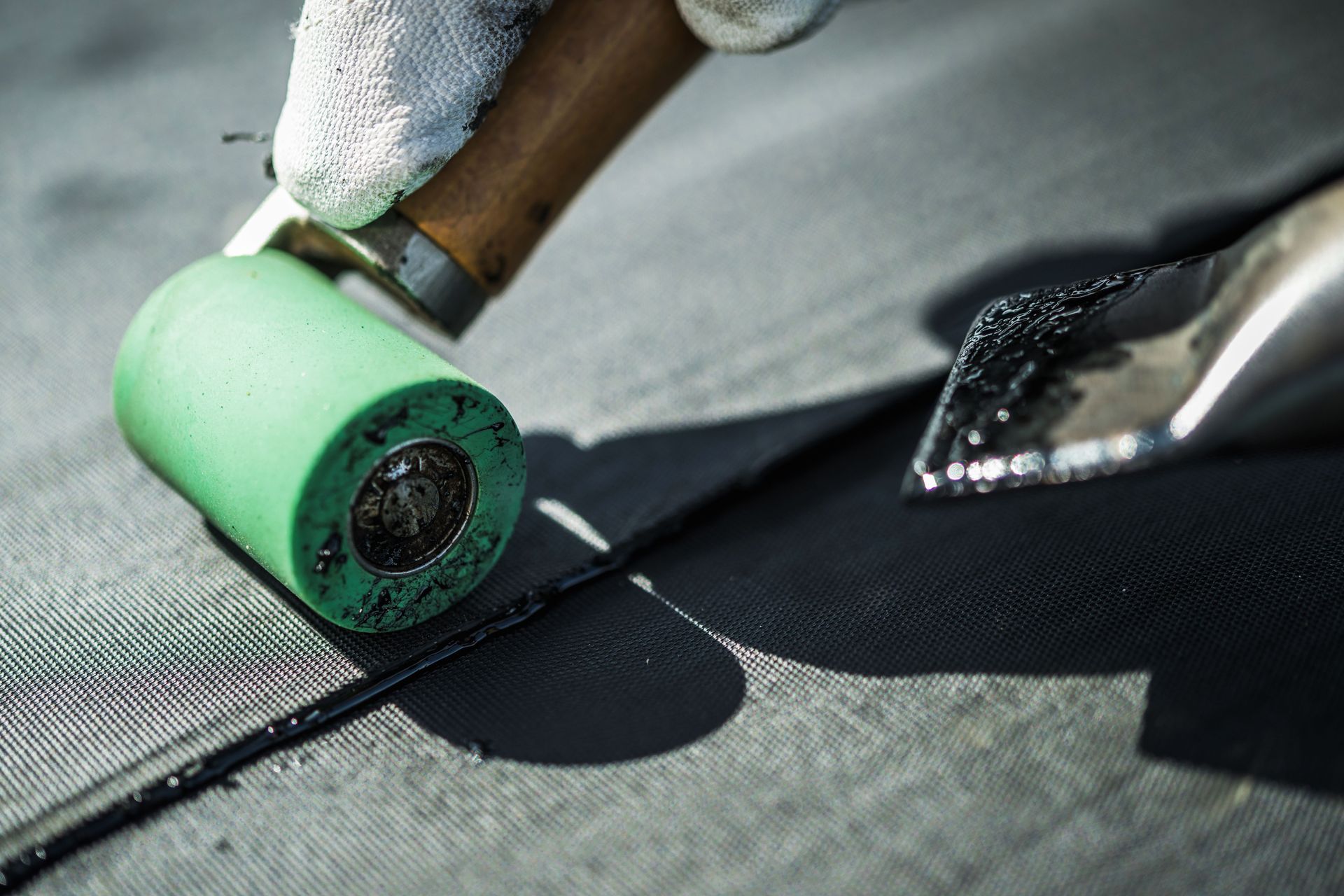 Person in gloves using a roller and tool to seal a dark roofing seam.