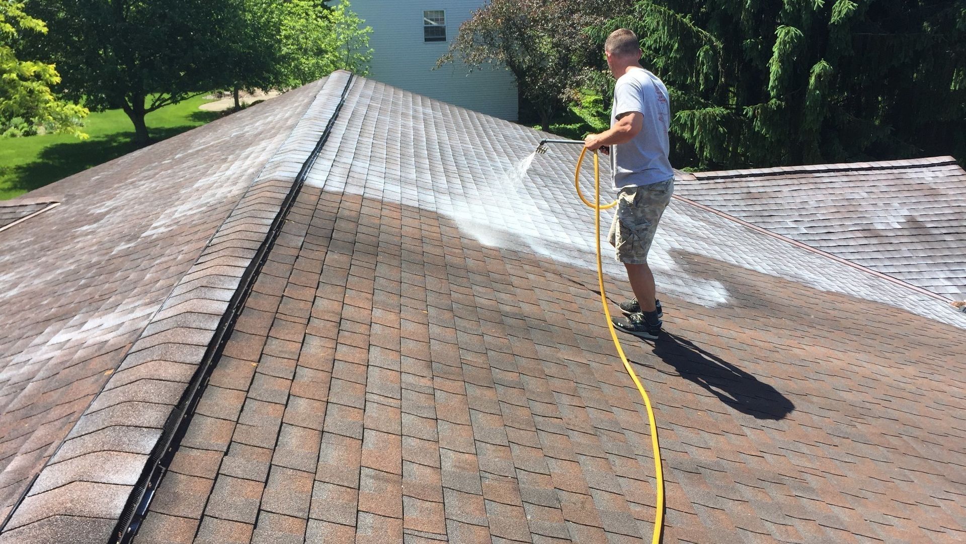 Man on roof spraying cleaning solution. Roof shingles are brown and covered in grime.