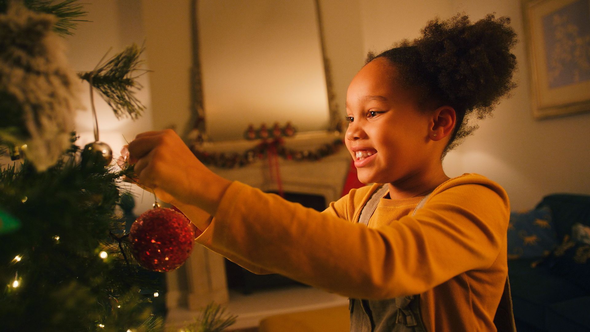 Girl decorating a Christmas tree with a red ornament in a warmly lit room.