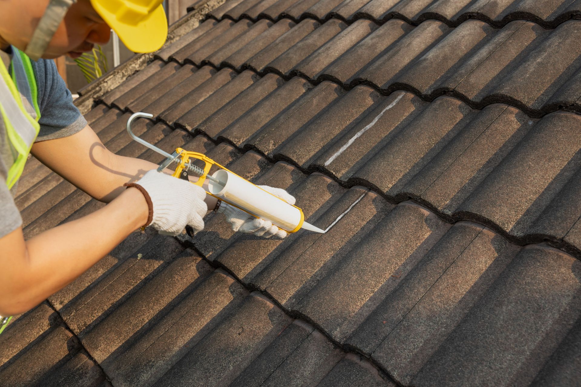 Person in a yellow hard hat and gloves applying sealant to roof tiles with a caulk gun.