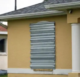 The front exterior of a tan stucco house featuring a large window completely covered by a vertical silver corrugated metal storm panel.