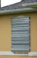 A beige house wall with a window boarded over by a gray metal panel near the roofline