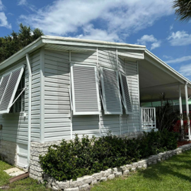 A light grey manufactured home with white siding, featuring adjustable louvered Bahama hurricane shutters propped open over the windows.