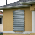 A beige house wall with a window boarded over by a gray metal panel near the roofline