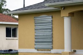 The front exterior of a tan stucco house featuring a large window completely covered by a vertical silver corrugated metal storm panel.