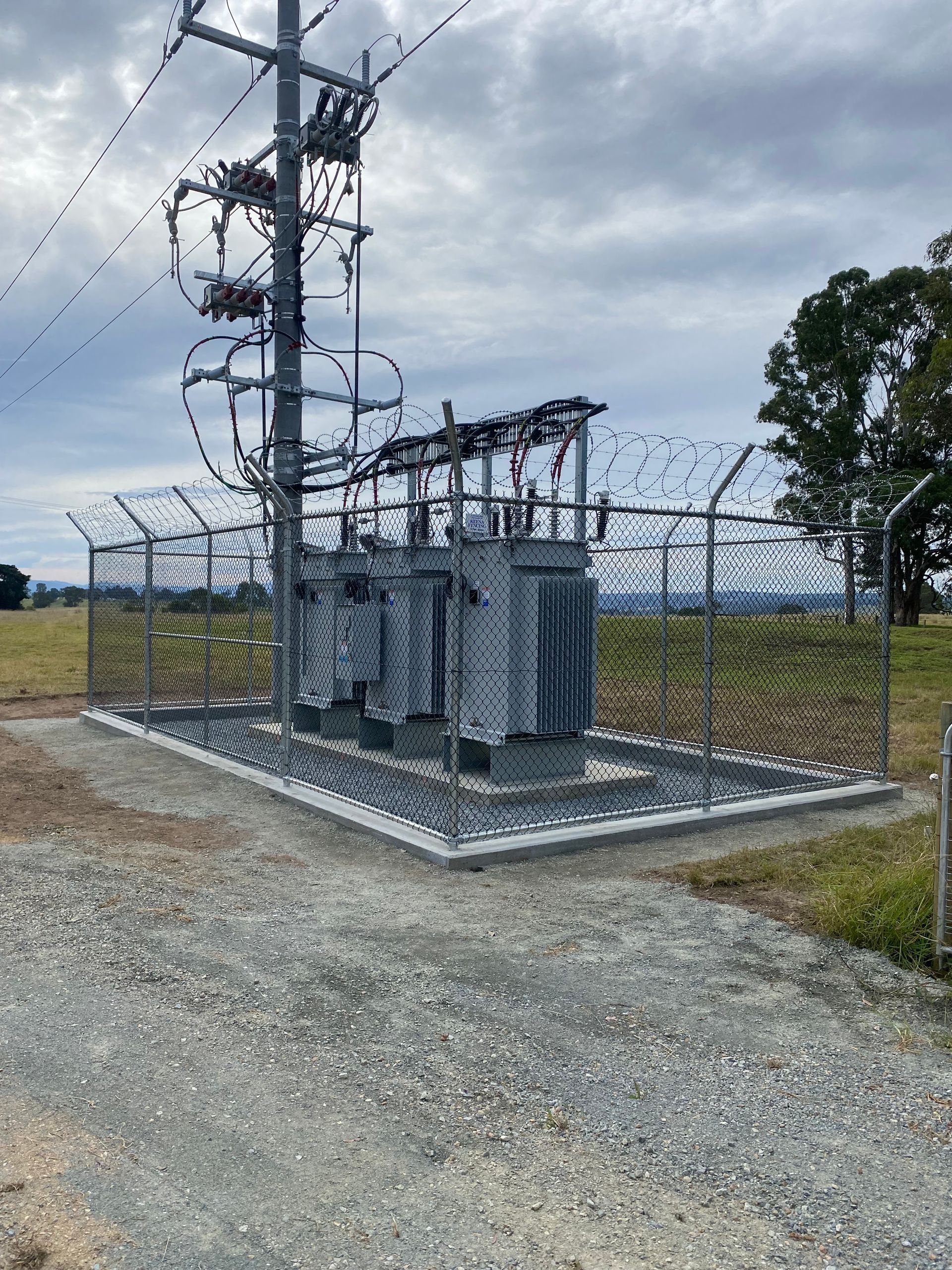 Power transformer station behind a chain link security fence, in a rural setting.