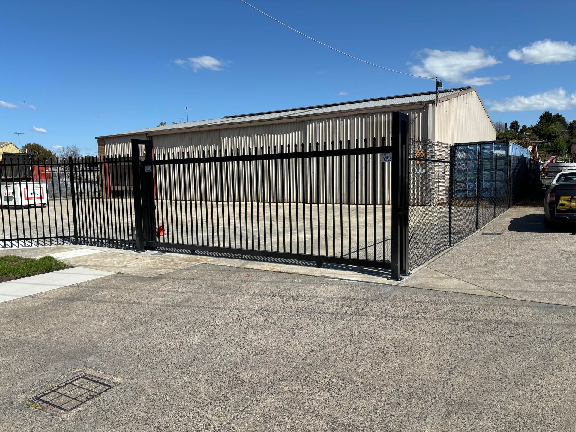 Black  palisade  sliding metal gates in front of a beige warehouse on a sunny day.