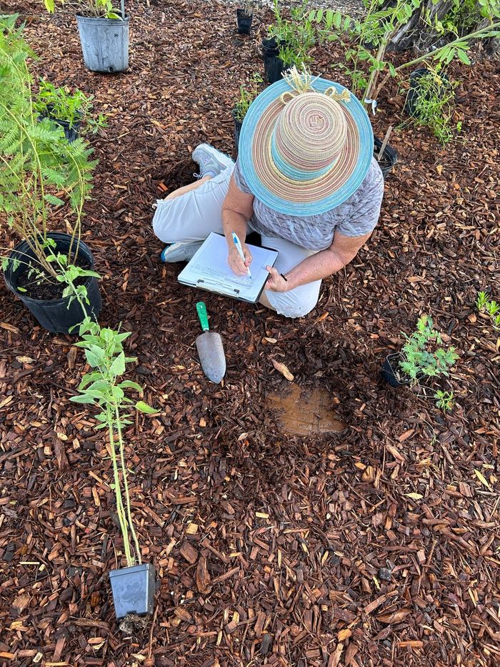 A woman is sitting on the ground in a garden writing in a notebook.