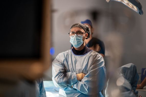 Un cirujano que lleva una mascarilla y una bata está de pie en una sala de operaciones con los brazos cruzados.