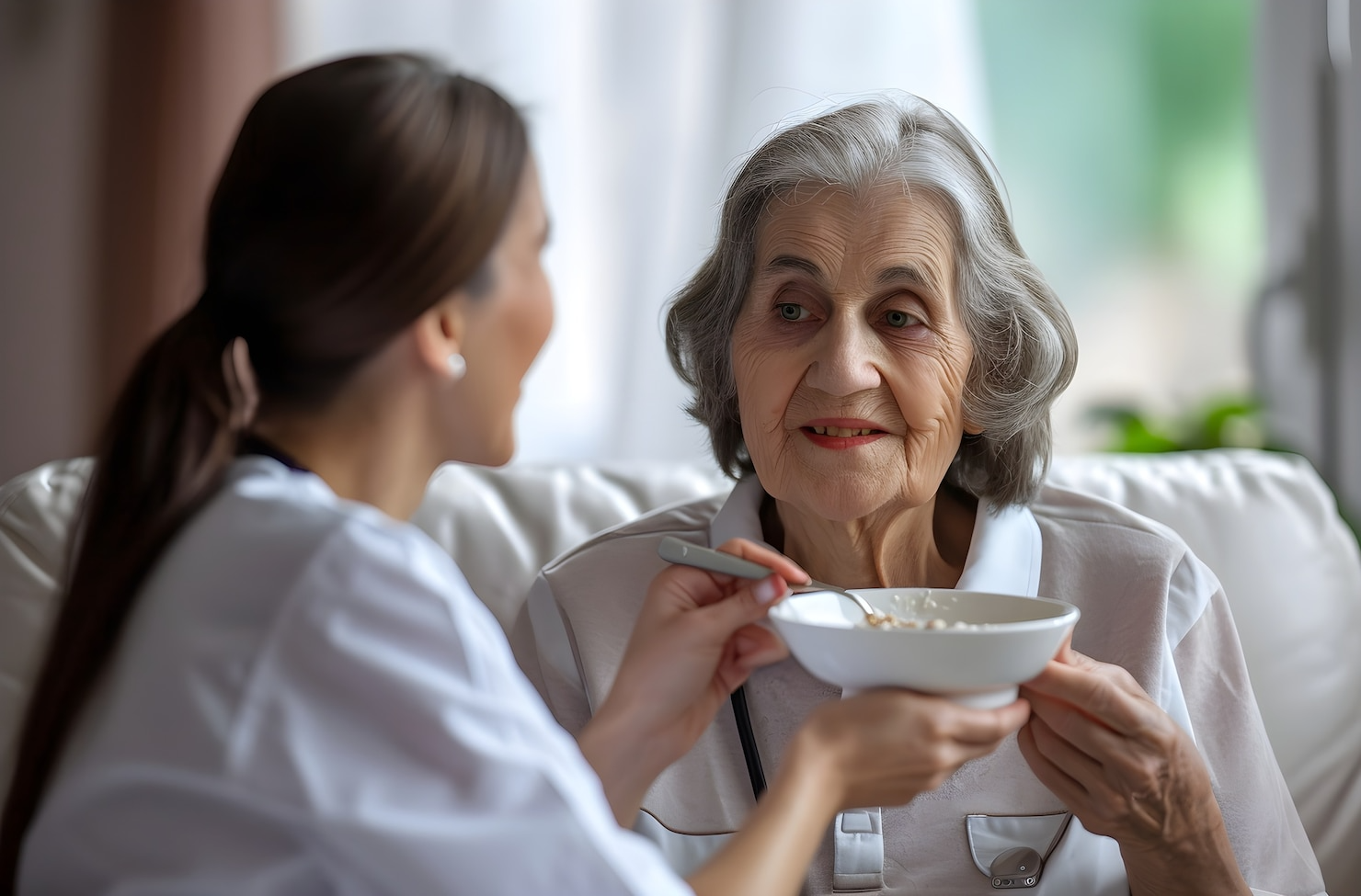 Caregiver feeding elderly woman oatmeal in a home setting; both smiling.