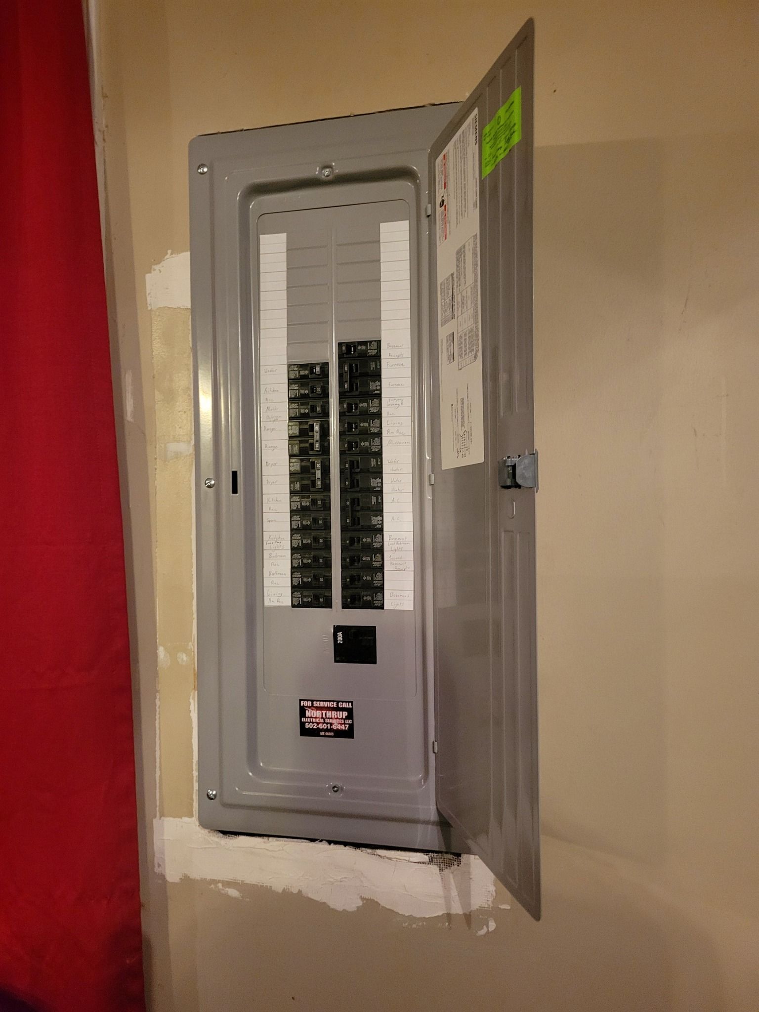 Electrical panel with open door, mounted on a wall. Gray metal, white wall, red fabric on the left.