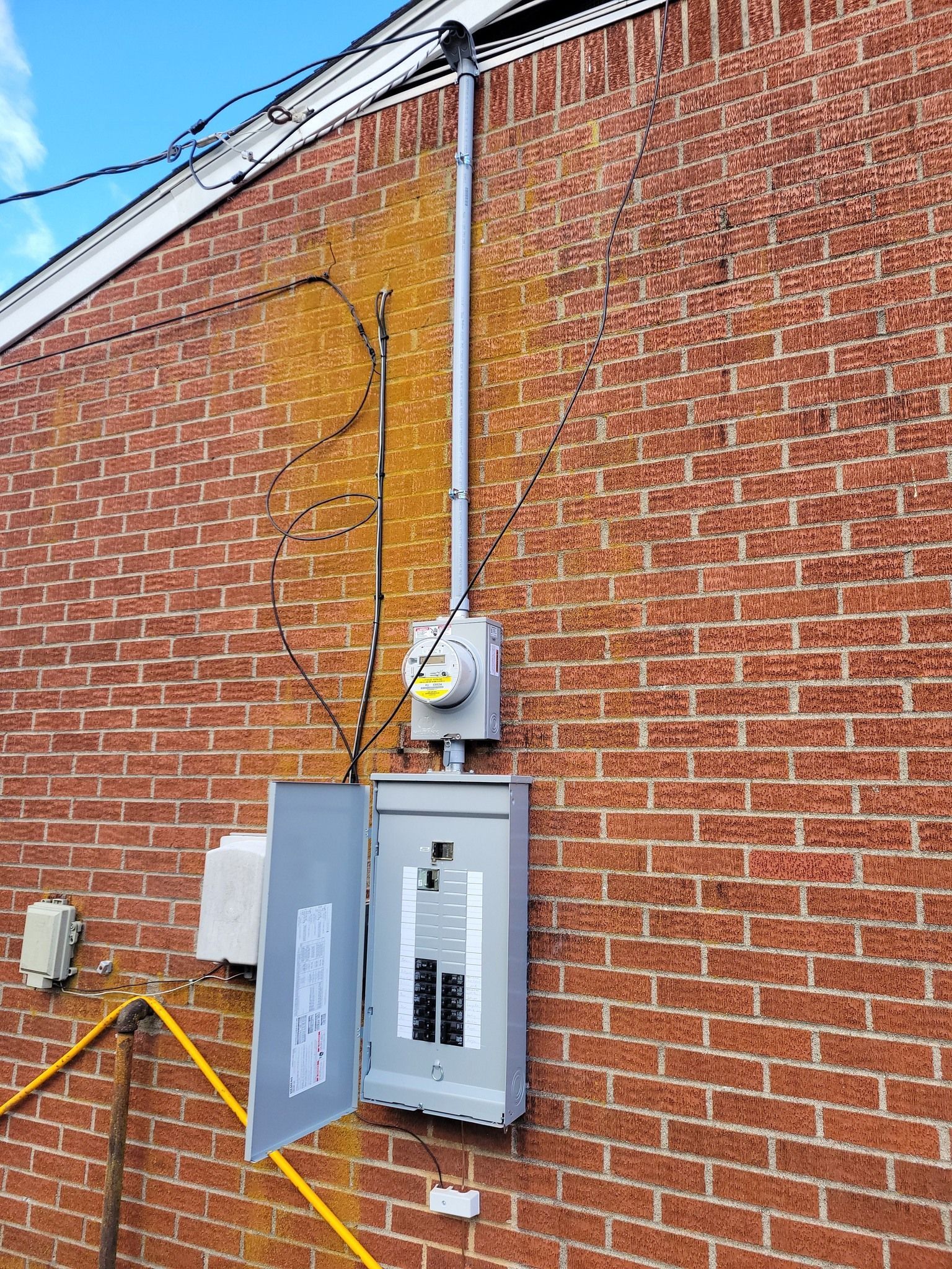 Electrical panel with meter on brick wall. Wires and conduit extend upwards. Open panel with circuit breakers.