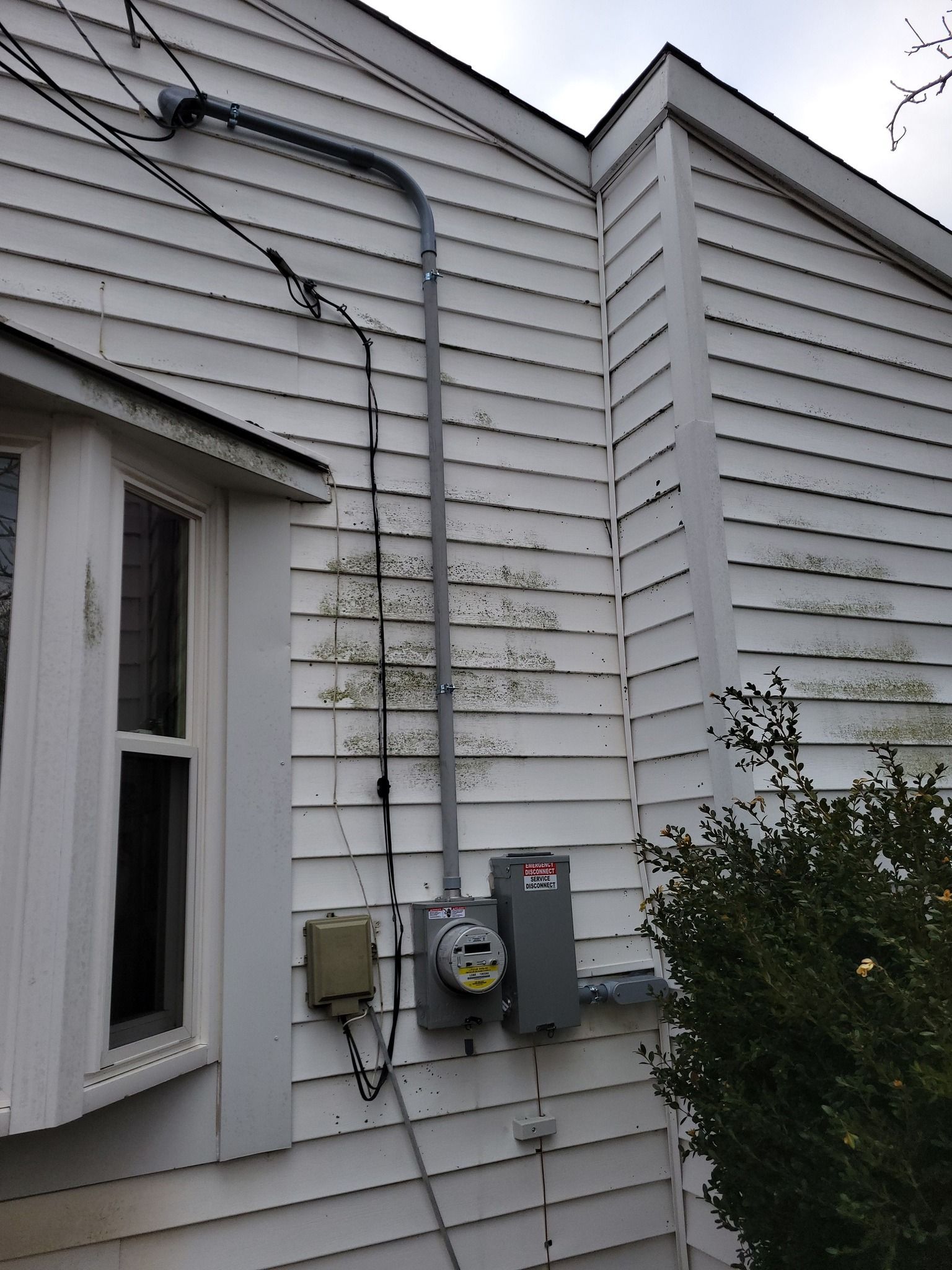Electrical panel and meter on a white house exterior with a conduit running up the wall.
