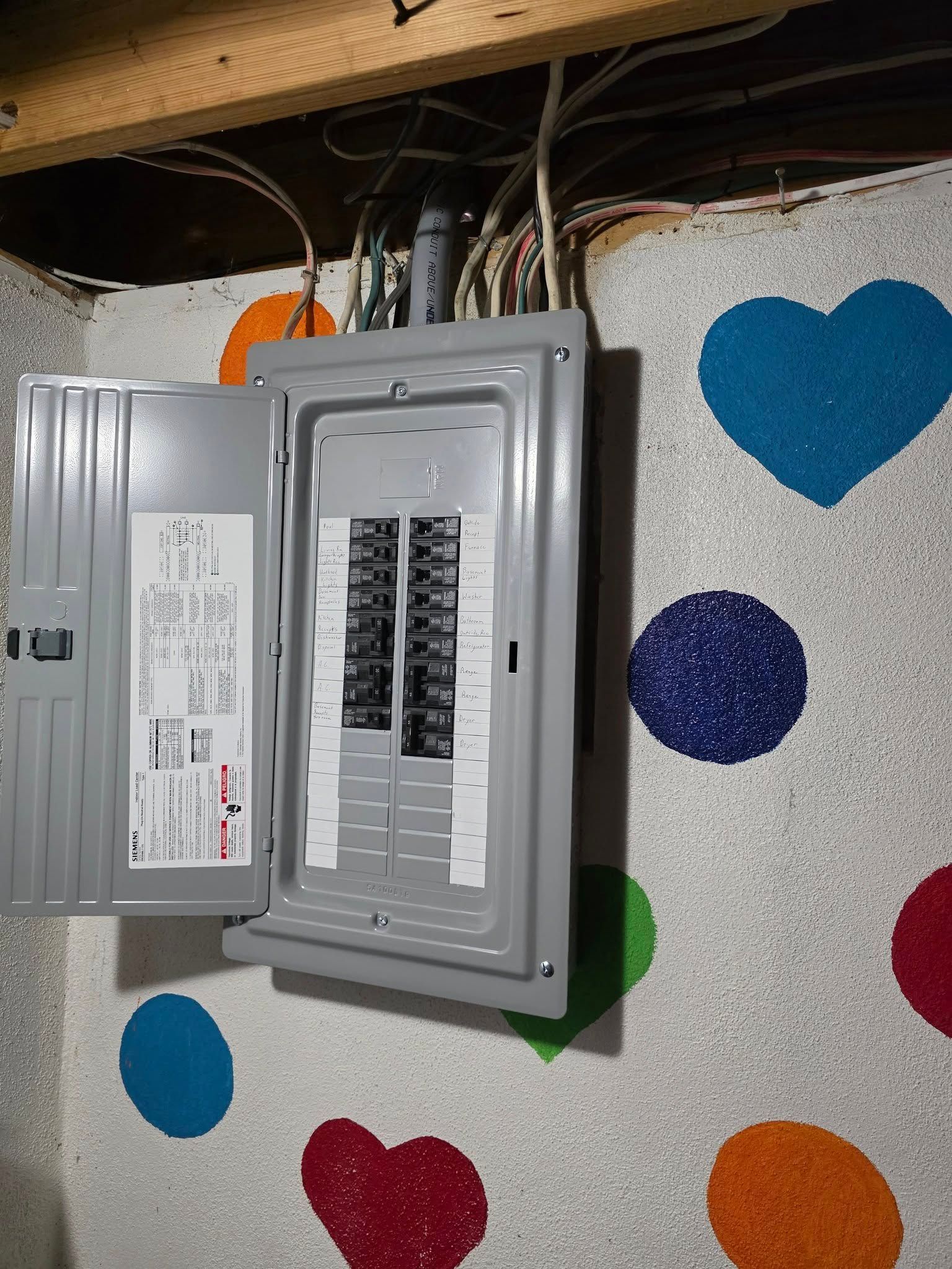 Open electrical panel on a wall with colorful heart and circle decorations. Wires are visible above.