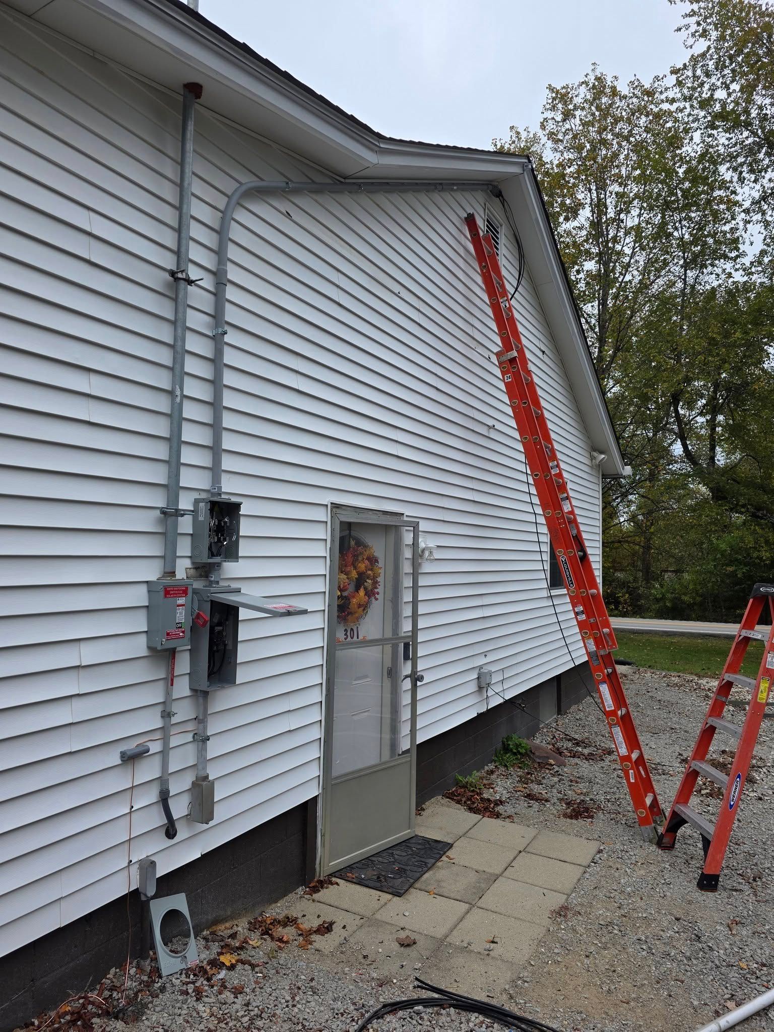 Electrical conduit and boxes on a white-sided building with a door, two red ladders, and a cloudy sky.