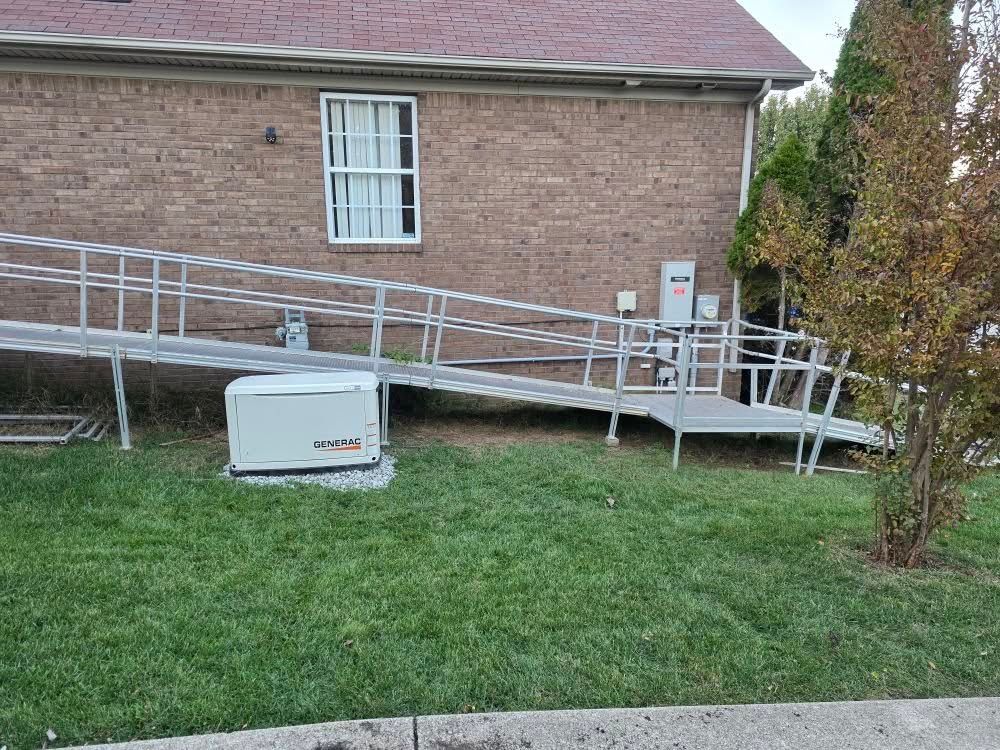 Aluminum ramp leads up to a house entrance. A generator sits on the ground. Brick building and green grass.