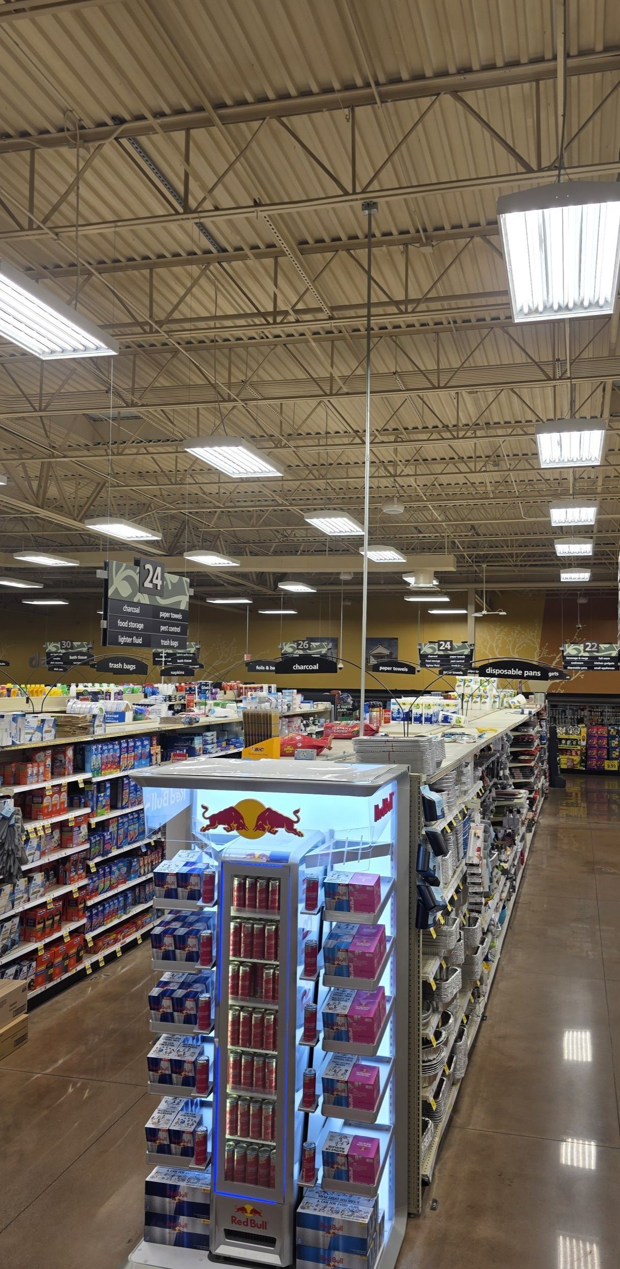 Interior view of a grocery store aisle with Red Bull display, other shelves of goods, and high ceiling.