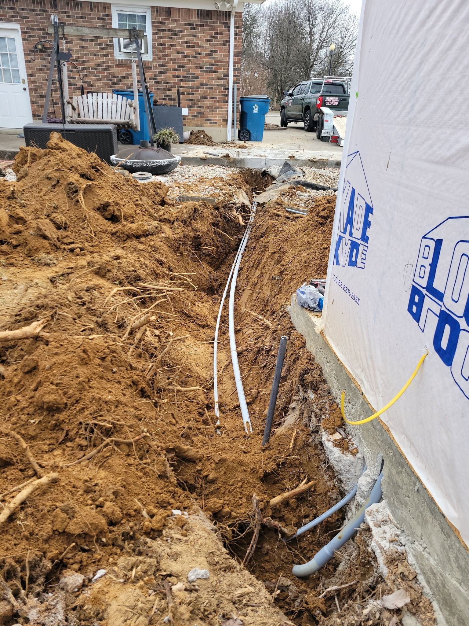 Construction site trench with buried pipes. A house's side is visible. Earth and tools are present.