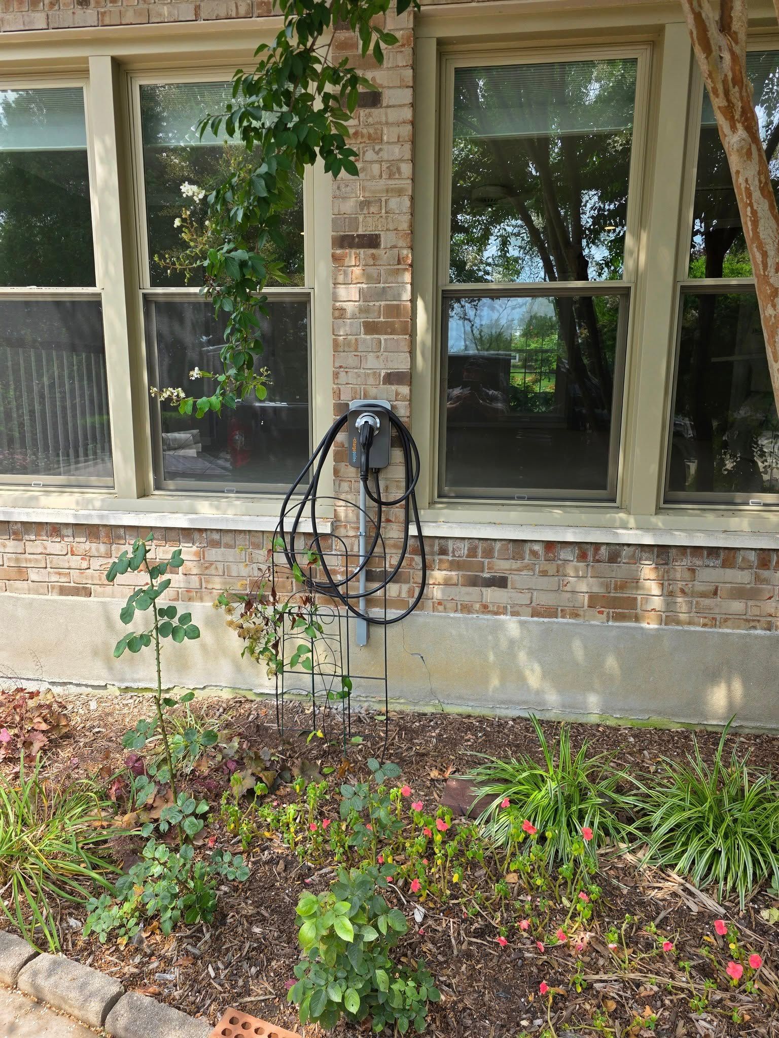 Electric car charger mounted on a brick wall, next to windows and landscaping.