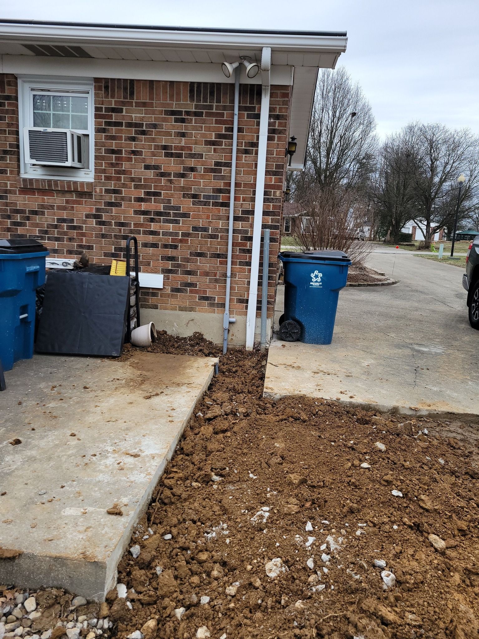Exterior of a brick building with a concrete sidewalk. Dirt is piled up along the side, next to a blue trash bin.