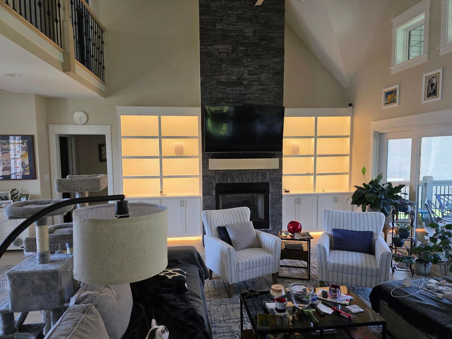 Living room with fireplace, built-in shelves, chairs, and large windows. Warm lighting and neutral colors.