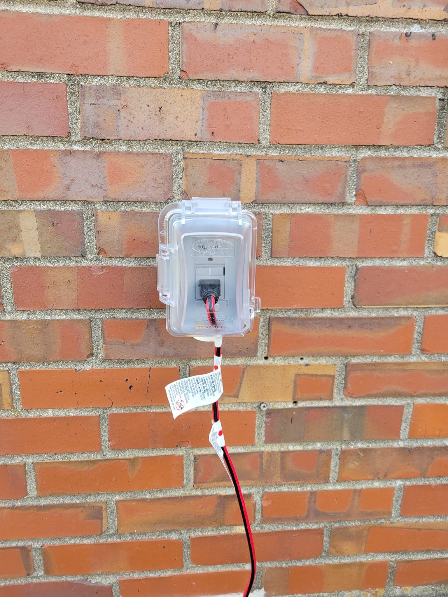 Clear plastic electrical box mounted on a red brick wall, with wires extending down.