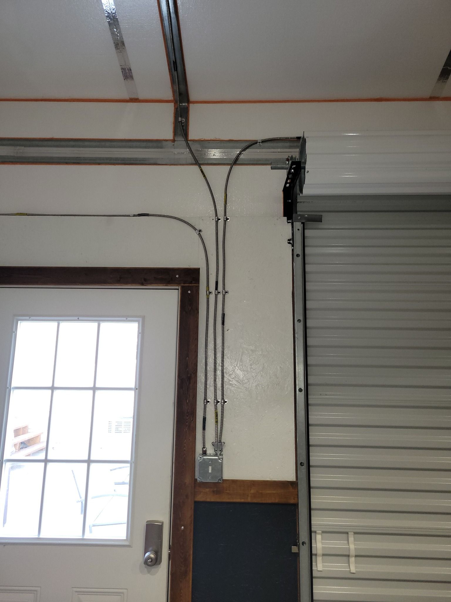 Electrical conduit and wiring run along a wall near a garage door and white door with window panes.