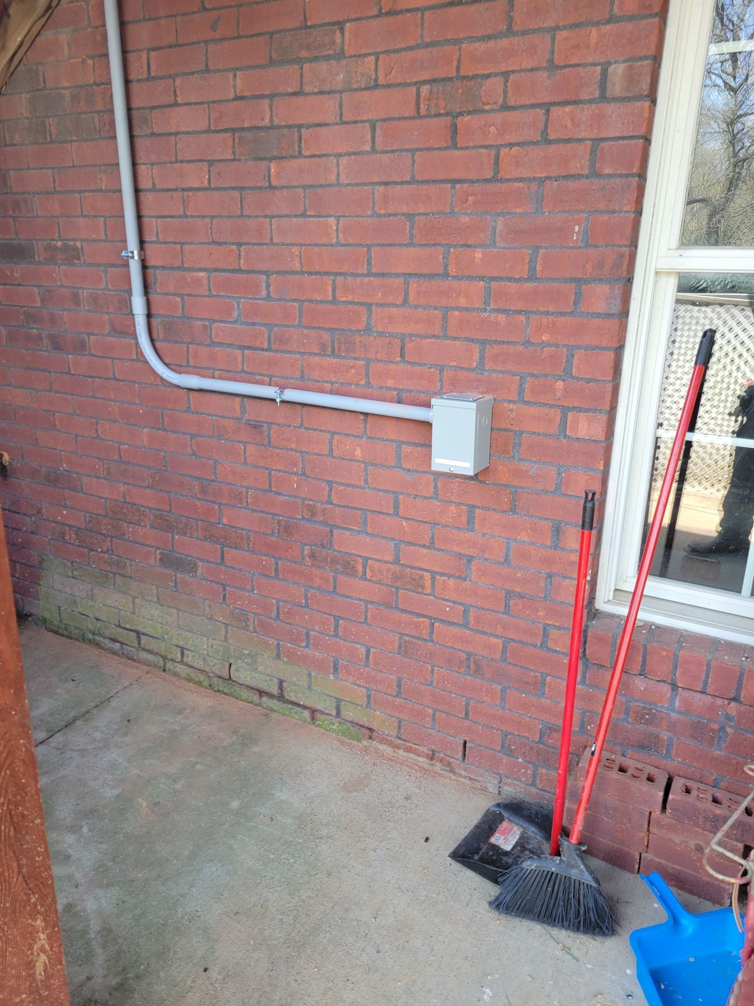 Red brick wall with electrical box, conduit, and window. Broom, dustpan, and debris on concrete.