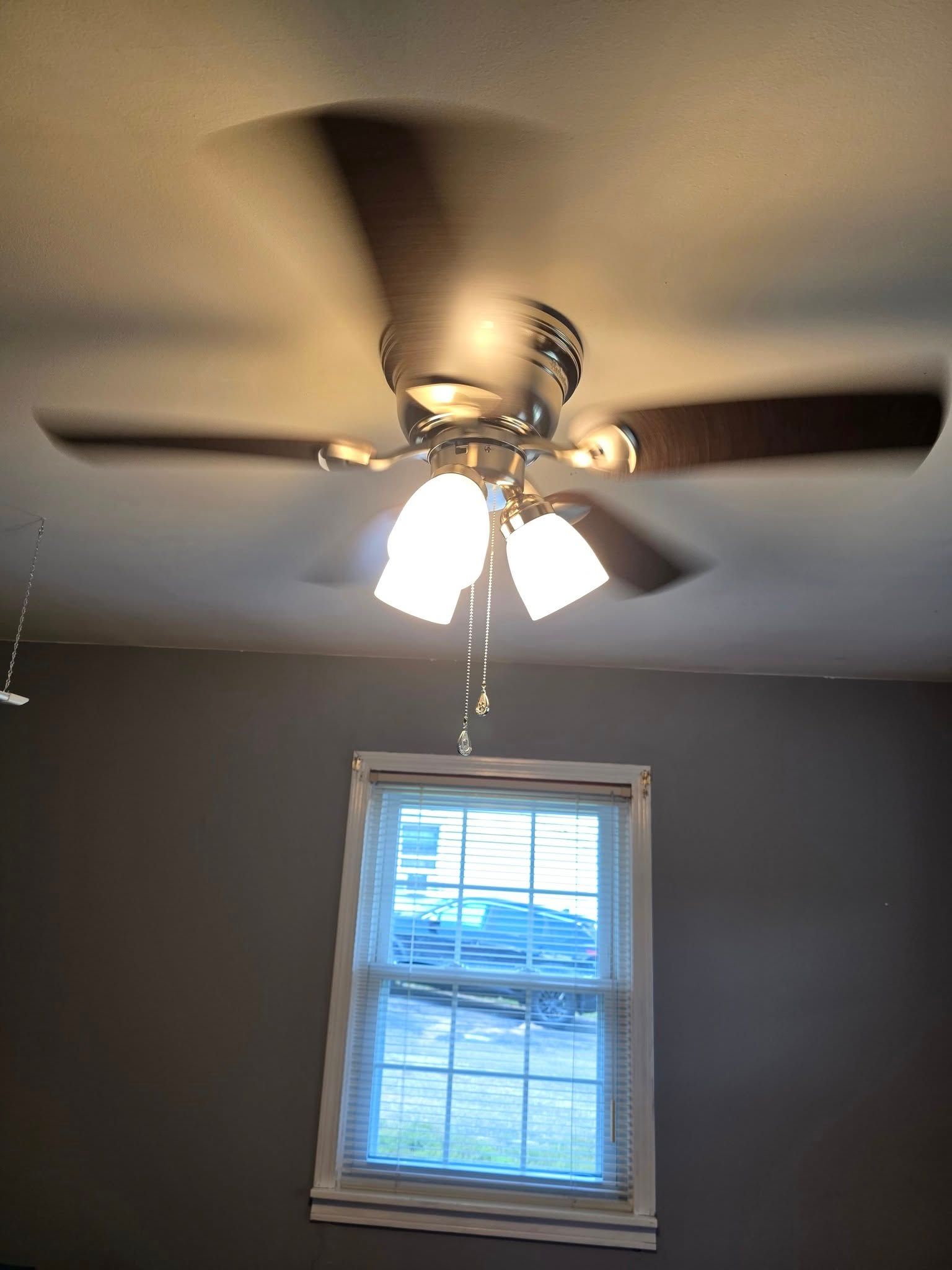 Ceiling fan with lights spinning above a small window in a room with gray walls.