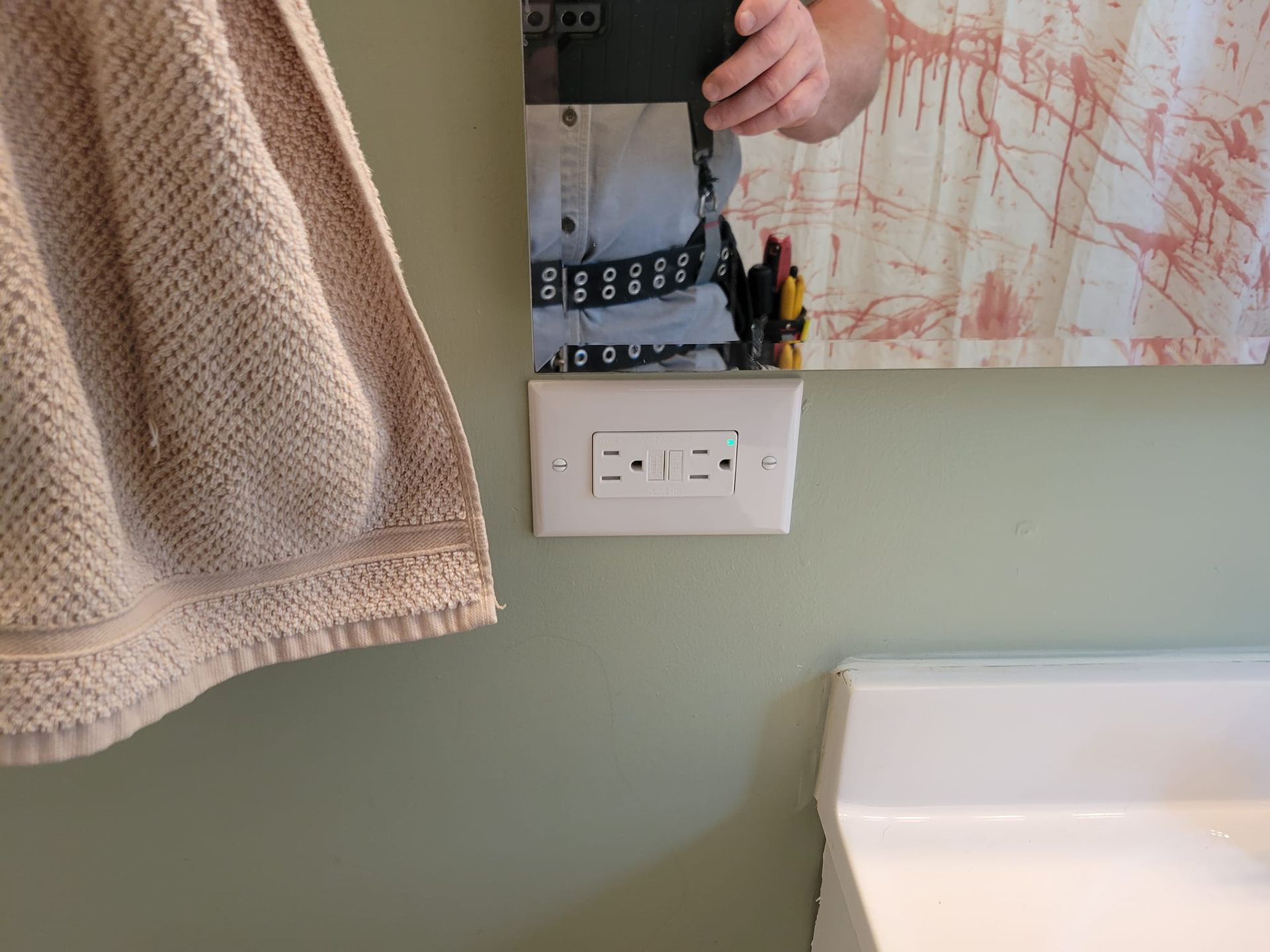 White outlet on a light green wall, near a hanging beige towel and a sink. A mirror reflects a person.