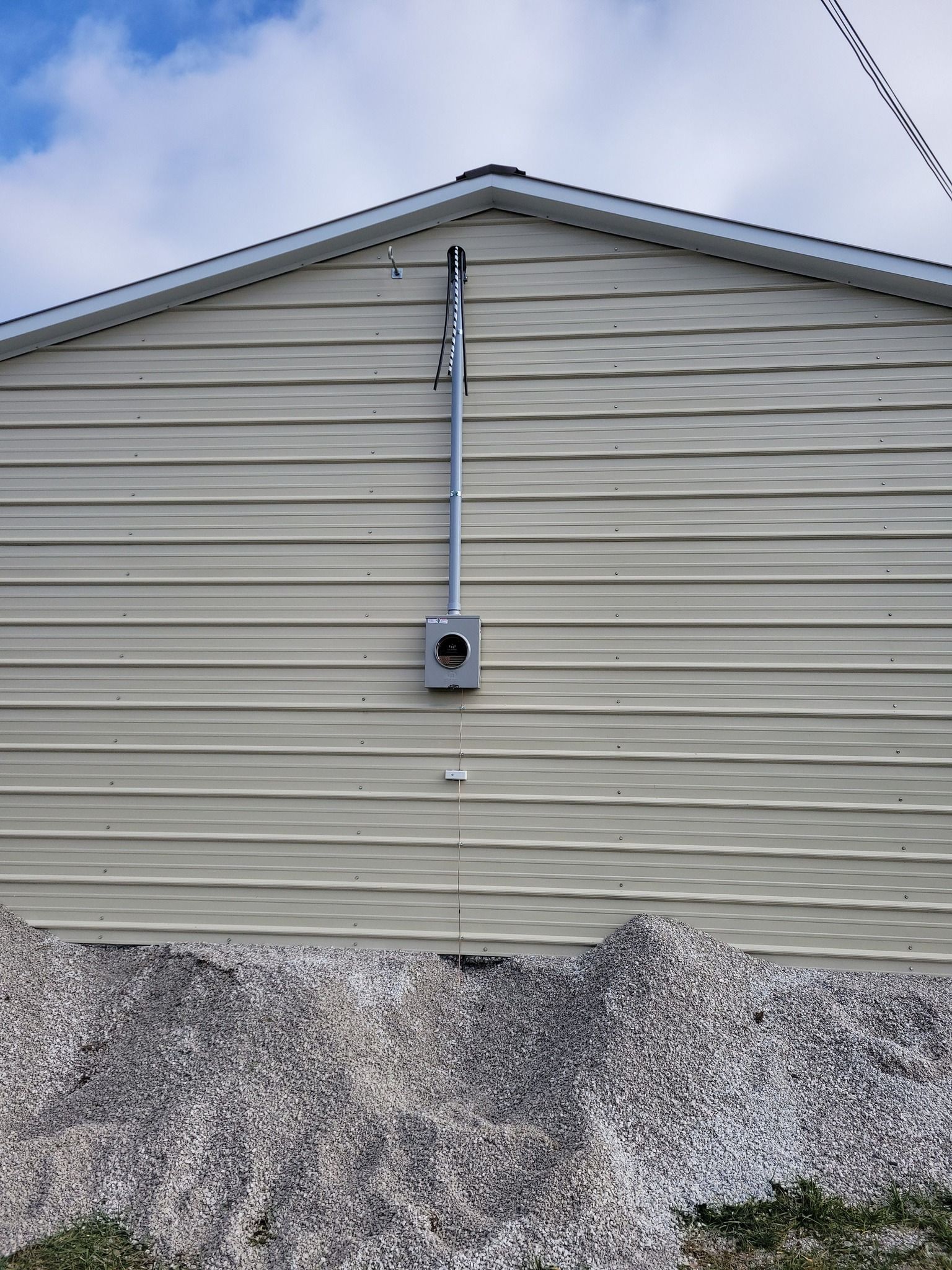 Electrical box and conduit mounted on beige siding of a building with gravel base and cloudy sky.