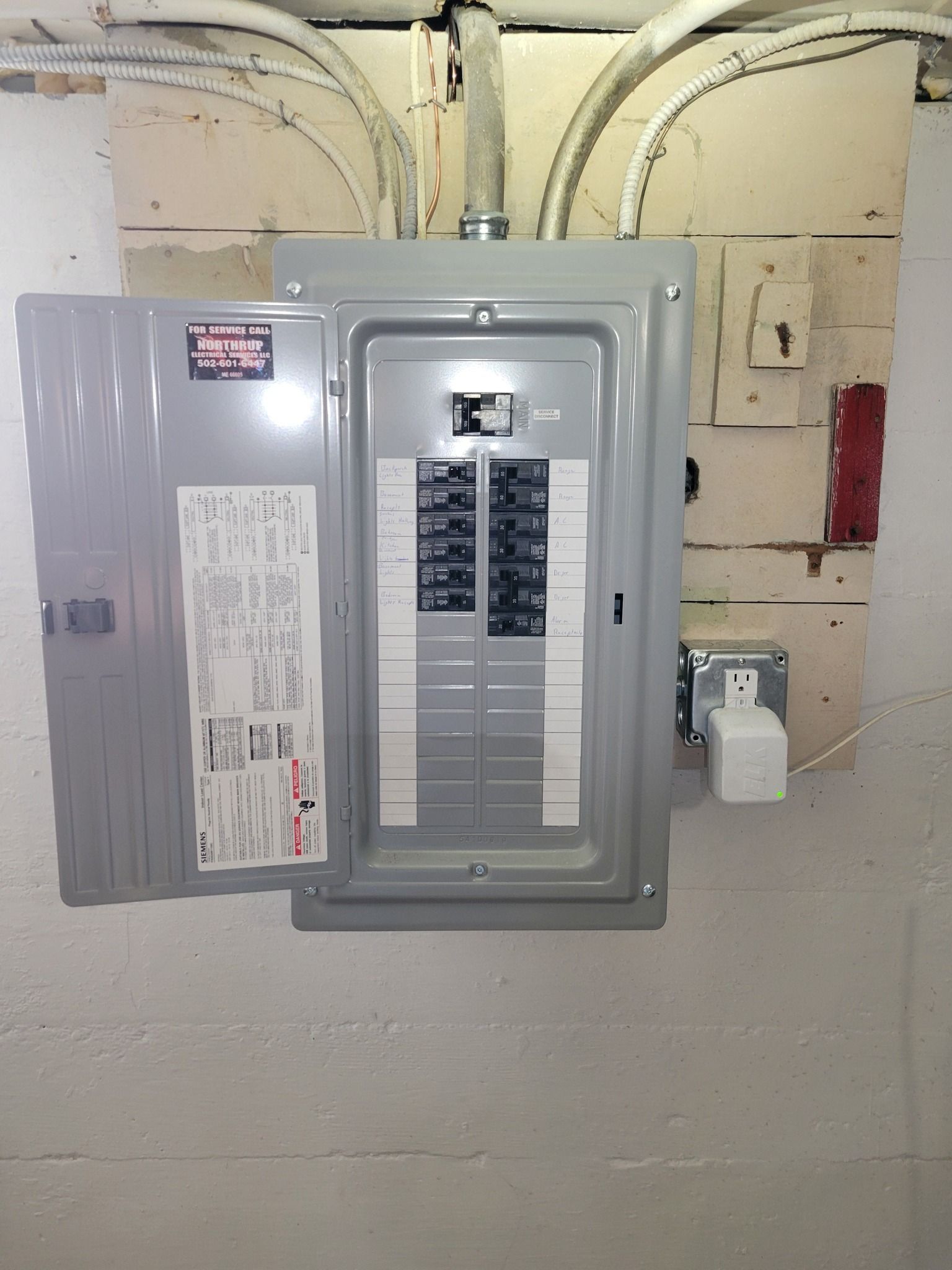 Electrical panel on a white wall, door open showing circuit breakers.  Outlet and wiring visible.