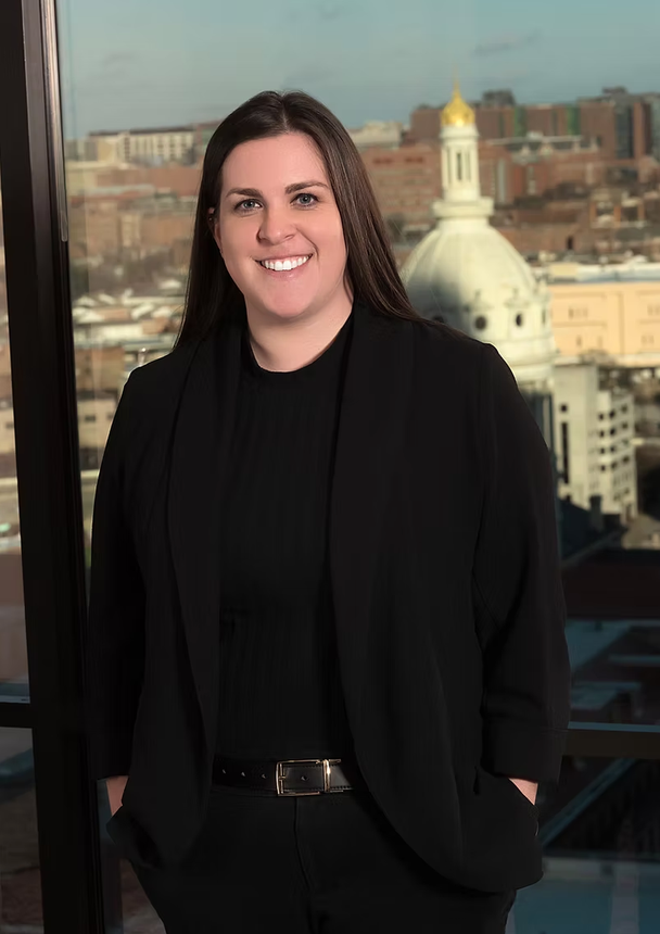 A woman in a black jacket is standing in front of a window with her hands in her pockets.