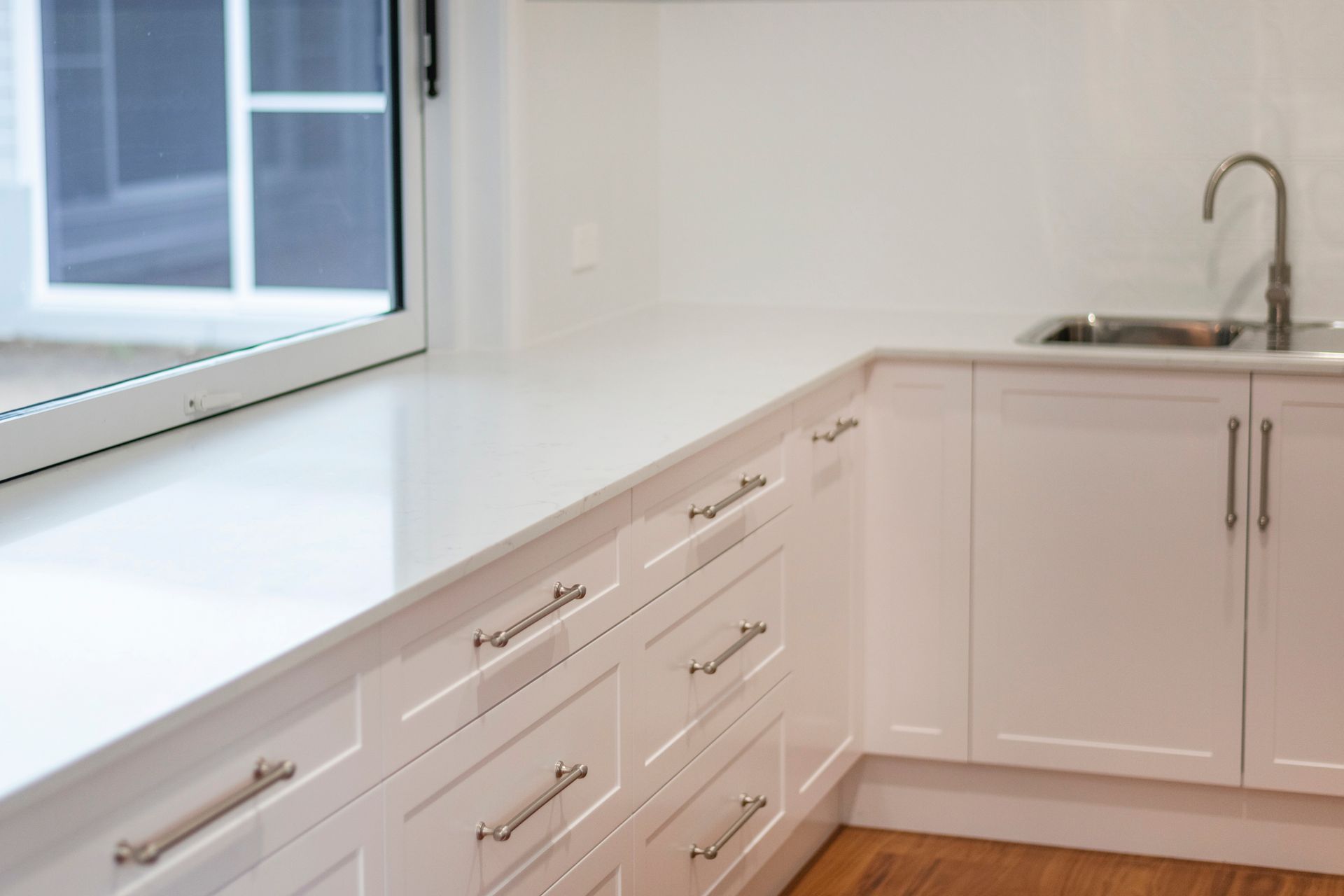 White kitchen cabinets and countertop with a stainless steel sink and faucet near a window.