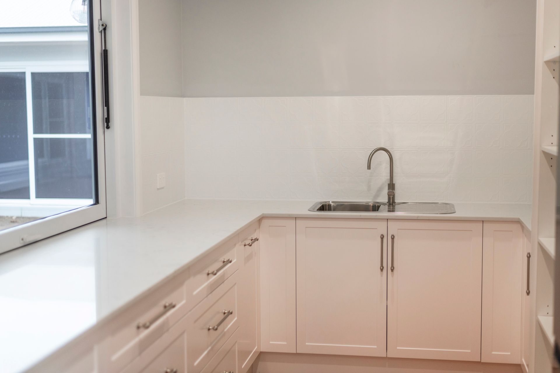 White cabinets and countertop in a kitchen area, with a sink under a tiled backsplash and a window.