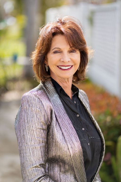 Smiling woman with dark hair wearing a silver sequined jacket and black shirt, standing outside near a white fence.