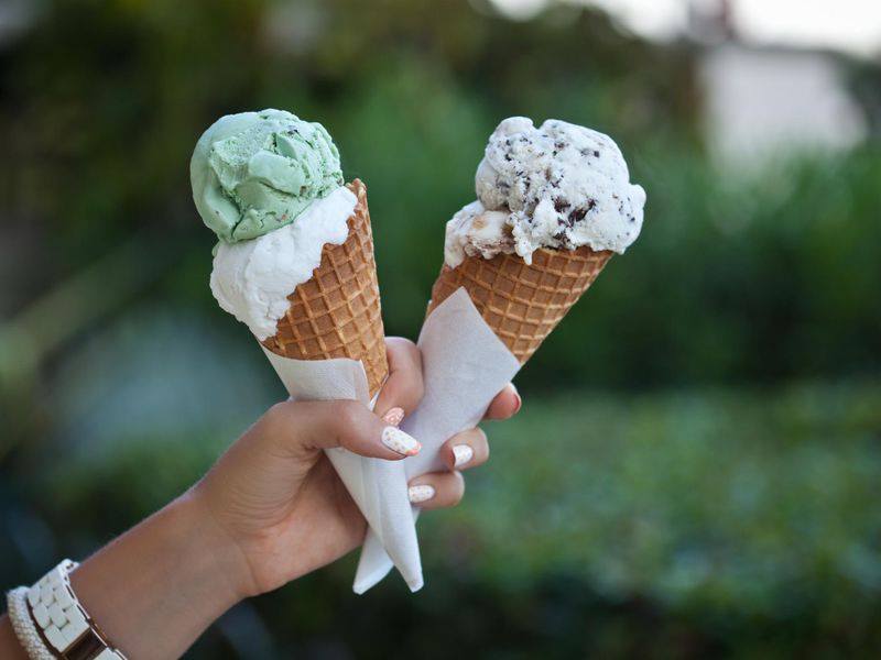 Hand holding two ice cream cones: mint and cookies-and-cream, against a blurred green background.