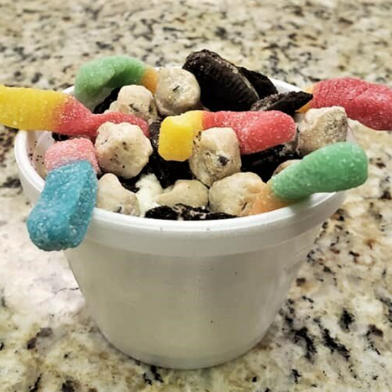 White cup filled with candy: Sour gummy worms, mini Oreos, and cookie dough bites.