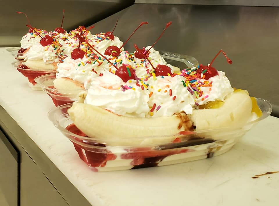 Banana splits with whipped cream, sprinkles, and cherries in plastic dishes.