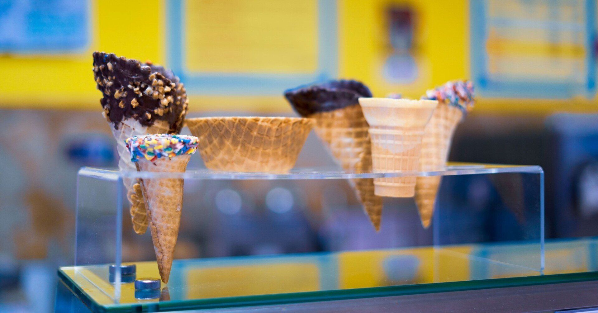 Ice cream cones on a glass display. Two cones are dipped in chocolate and sprinkles, others are empty.