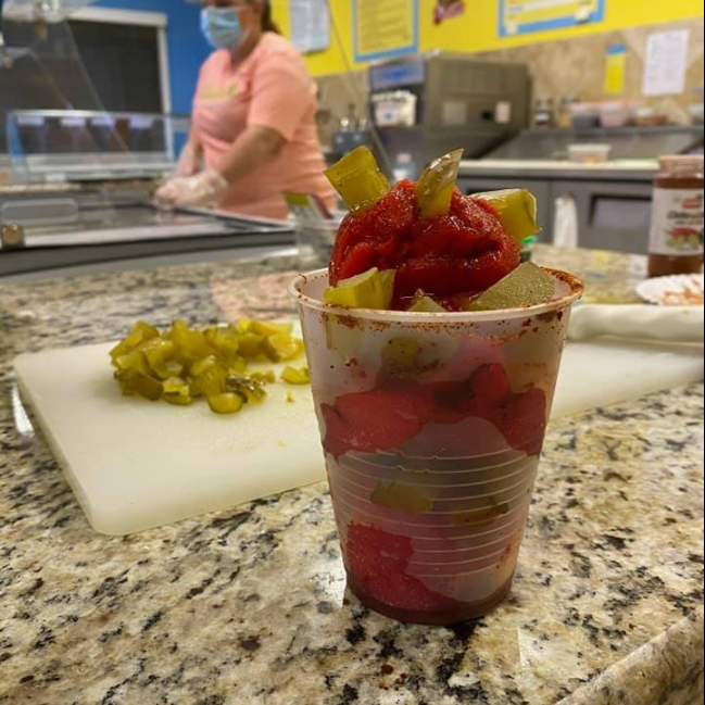 A cup with fruit, pickles, and a red sauce on a counter with a person in background.