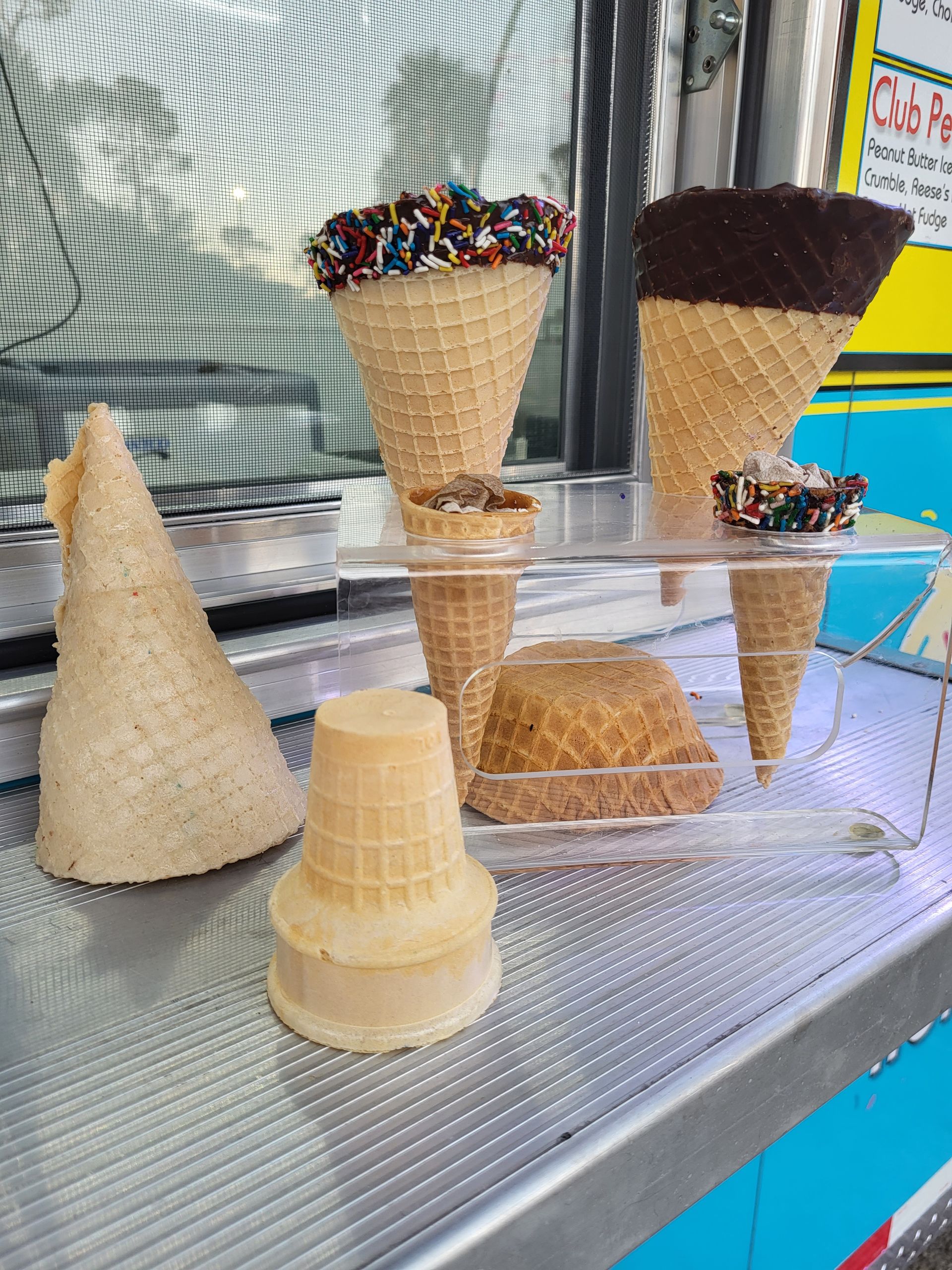 Assortment of waffle cones: plain, dipped in chocolate, and with sprinkles on a display, likely at an ice cream shop.