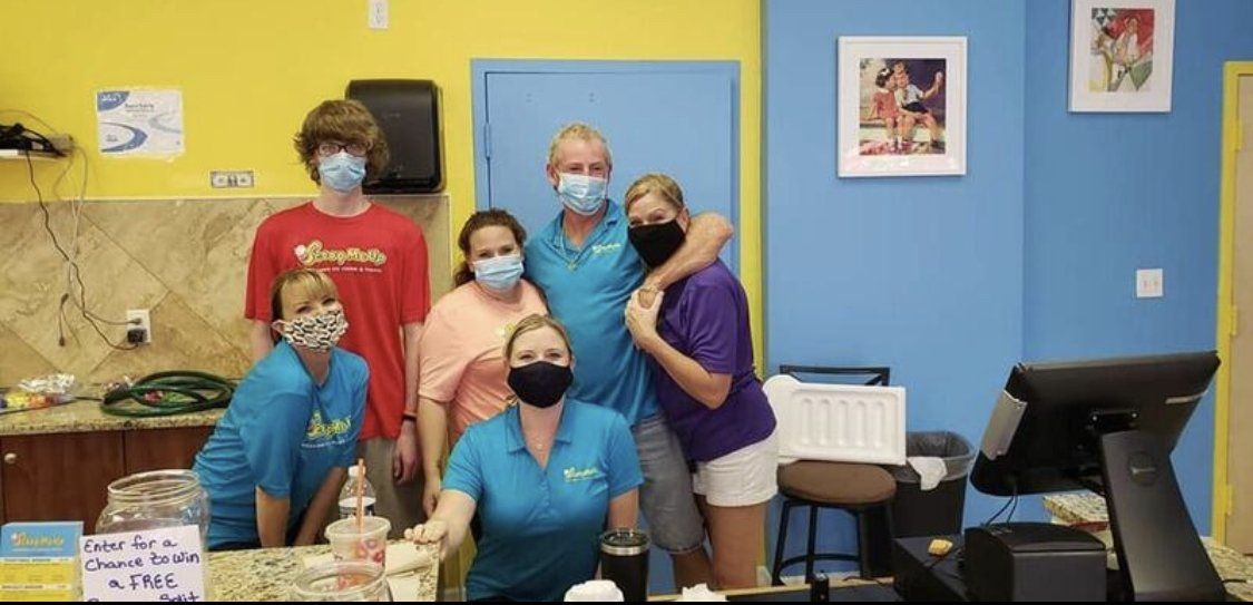 Group of people wearing masks posing for a photo near a counter, blue and yellow walls.