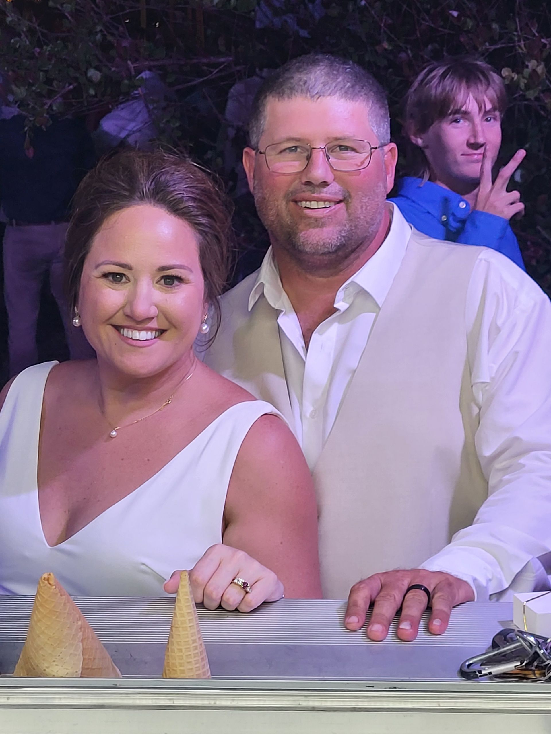 Couple smiling at the camera, woman in white dress. Man in vest, background of ice cream cones and a young person.